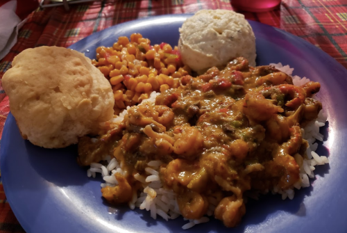 A blue plate with rice topped with shrimp stew, corn, and two biscuits, set on a red plaid tablecloth.