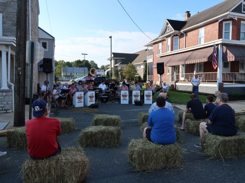 A community band performs outdoors while people sit on hay bales, enjoying the music in a neighborhood setting.
