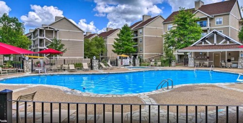 A sunny pool area with lounge chairs, surrounded by trees and apartment buildings, featuring red umbrellas and a stone path.