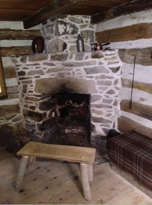 A rustic stone fireplace in a log cabin, with a wooden bench and a plaid-covered box nearby.