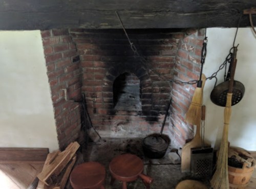 A rustic brick fireplace with a darkened chimney, surrounded by wooden stools and cleaning tools.