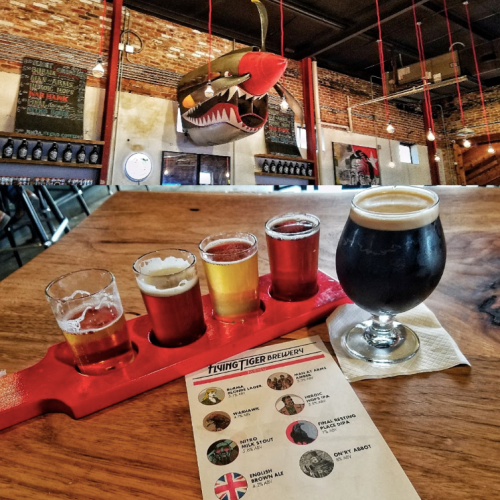 A wooden table with a flight of four beer samples and a pint, alongside a menu in a brewery with a shark mural.