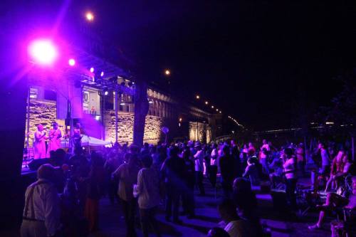 A lively outdoor night event with a crowd enjoying a performance under purple stage lights.