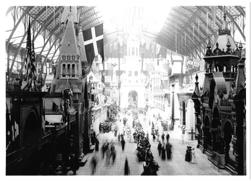 A historic black-and-white photo of a bustling indoor market with ornate architecture and flags hanging from the ceiling.