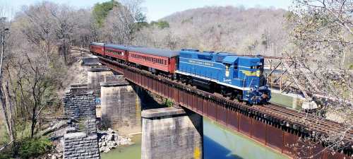 A blue locomotive pulls red passenger cars across a bridge over a river, surrounded by trees and hills.