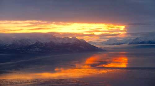 A stunning sunset over snow-capped mountains, reflecting golden hues on a calm body of water.