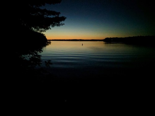 A serene lake at dusk, with gentle ripples and a silhouette of a person standing in the water against a colorful horizon.