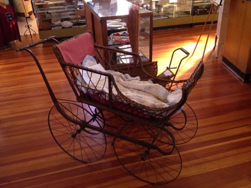 A vintage baby carriage with a quilted blanket, displayed on a wooden floor in a museum setting.
