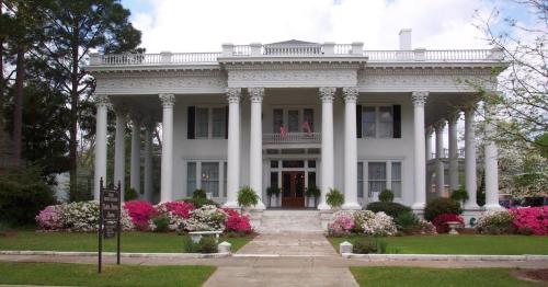 A grand white mansion with columns, surrounded by colorful flowers and greenery, under a partly cloudy sky.