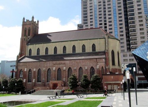 Historic church building with a tall tower, surrounded by modern skyscrapers and green landscaping.