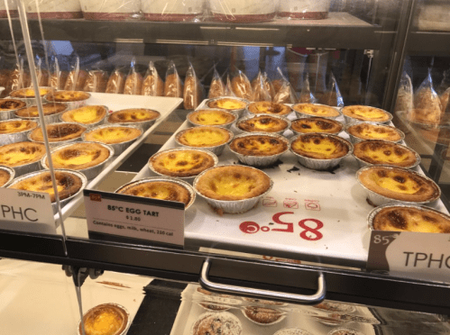 A display case filled with golden-brown egg tarts in individual aluminum cups, labeled with prices and calorie information.