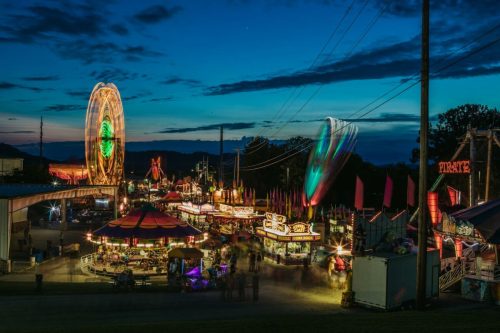A vibrant carnival scene at dusk, featuring a Ferris wheel, colorful rides, and illuminated food stalls.