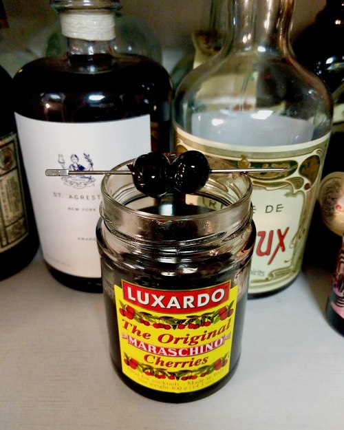 A jar of Luxardo maraschino cherries with a cherry on a pick, surrounded by bottles of spirits in the background.