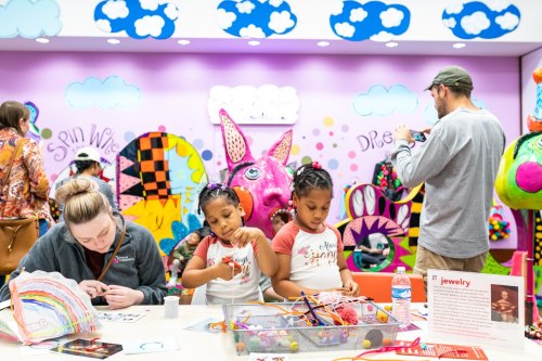 Two girls create art at a colorful table while an adult works in the background, surrounded by vibrant decorations.