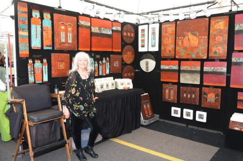 A woman stands in front of her art display featuring colorful paintings and framed pieces at an outdoor market.