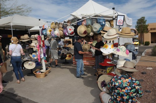 A market stall displays a variety of hats, with customers browsing and trying on different styles under a sunny sky.