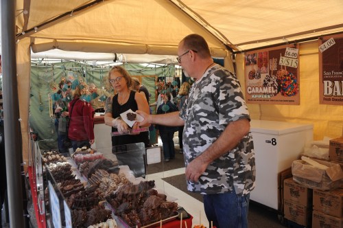 A vendor and a customer interact at a market stall filled with various chocolate treats and caramel apples.