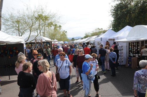 A bustling outdoor market with numerous tents, people browsing, and a backdrop of trees and mountains.
