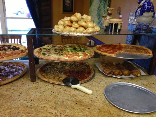 A display of various pizzas and baked goods on a counter, with a pizza cutter and empty tray in front.