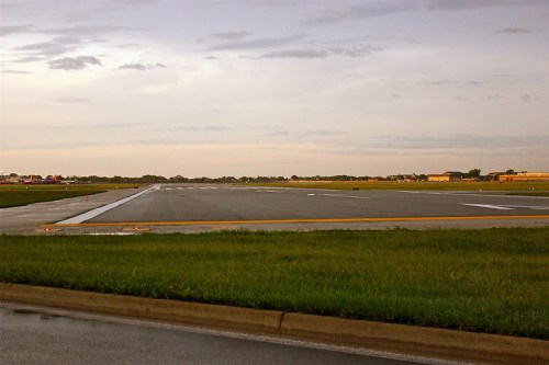 A view of an empty airport runway with wet pavement and a cloudy sky in the background.