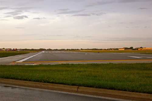 A view of an empty airport runway with wet pavement and a cloudy sky in the background.