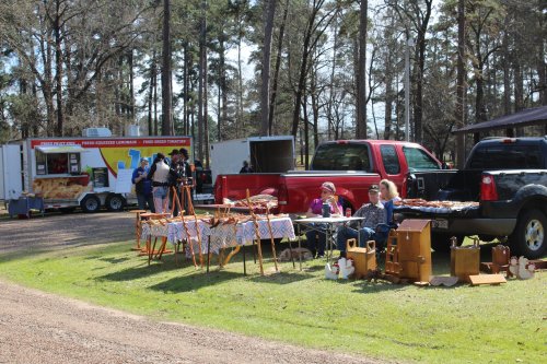 A sunny outdoor market scene with vendors selling wooden items, food trucks, and people browsing among trees.