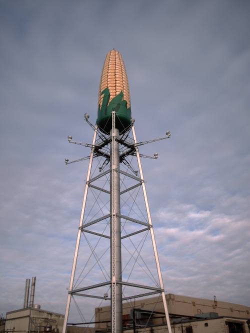 A tall tower shaped like an ear of corn, standing against a cloudy sky, with industrial buildings in the background.