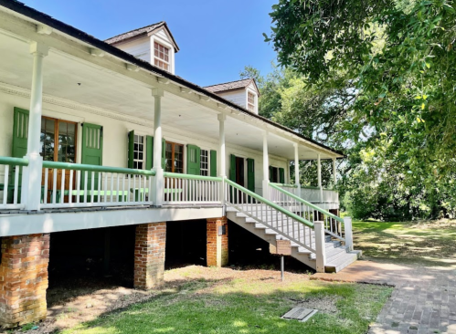 A historic house with green shutters and a porch, surrounded by trees and a grassy area under a clear blue sky.