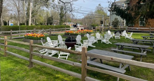 A grassy area with white Adirondack chairs, picnic tables, and flower beds, surrounded by a wooden fence.