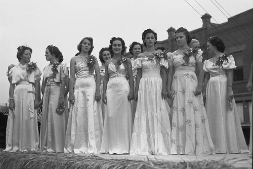 A group of young women in elegant dresses stand together on a stage, smiling at an outdoor event.