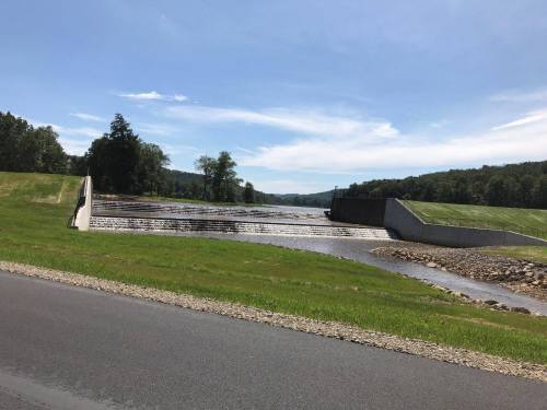 A serene landscape featuring a river flowing over a dam, surrounded by green grass and trees under a clear blue sky.