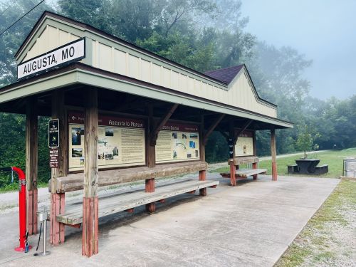 A small train station shelter in Augusta, MO, with benches and informational displays, surrounded by trees and fog.