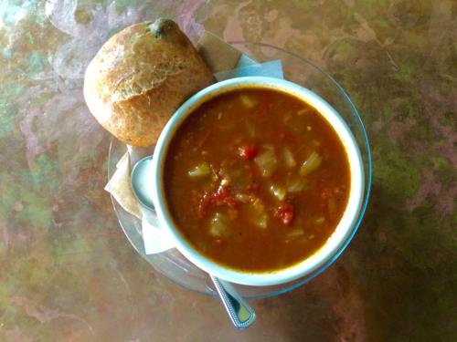 A bowl of hearty soup with vegetables, served with a small roll on a plate.
