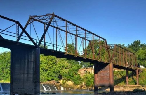 An old, rusted truss bridge spans a river, surrounded by lush greenery and a small waterfall in the background.
