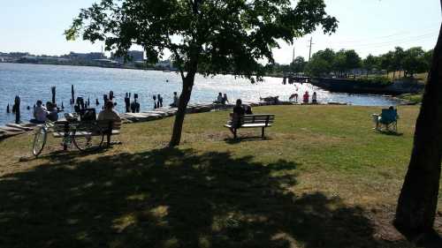 A sunny riverside park with people sitting on benches, enjoying the view, and a few boats on the water.