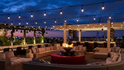 Rooftop lounge with cozy seating, a fire pit, and string lights, overlooking a city skyline at dusk.