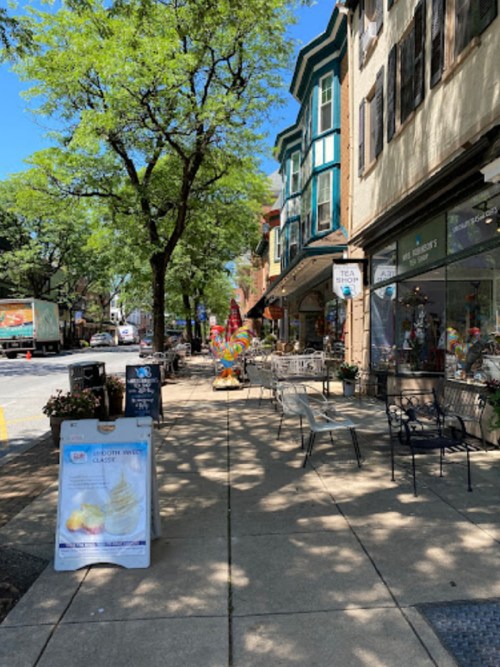 A sunny street scene with shops, outdoor seating, and colorful decorations along the sidewalk.