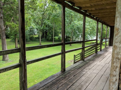 A wooden porch overlooking a green lawn surrounded by trees, with a peaceful, natural setting.