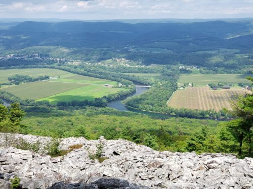 A panoramic view of lush green valleys and a winding river, framed by rocky terrain and distant hills under a blue sky.