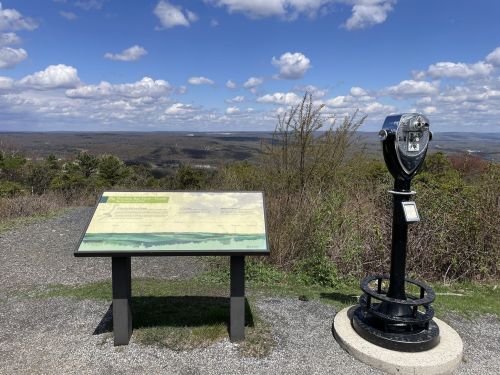 A scenic overlook with a viewfinder and informational sign, surrounded by greenery and a blue sky with clouds.