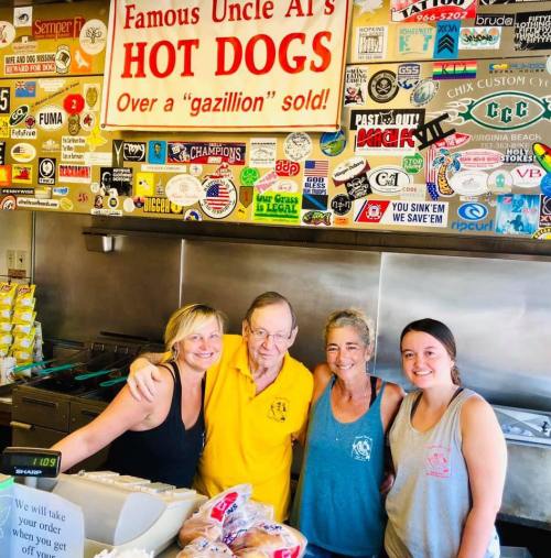 A group of four people, including an older man in a yellow shirt, stand behind a hot dog stand covered in stickers.