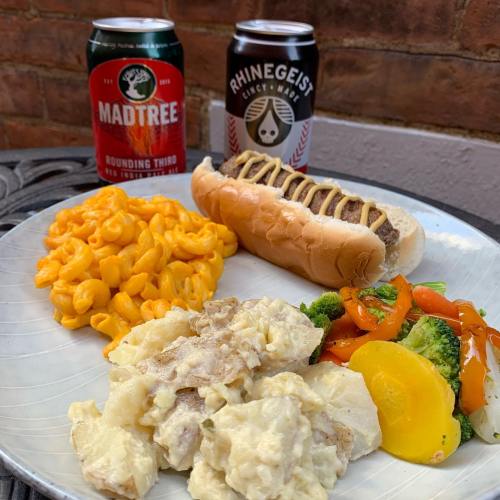 A plate with macaroni and cheese, potato salad, mixed vegetables, and a hot dog, alongside two cans of beer.