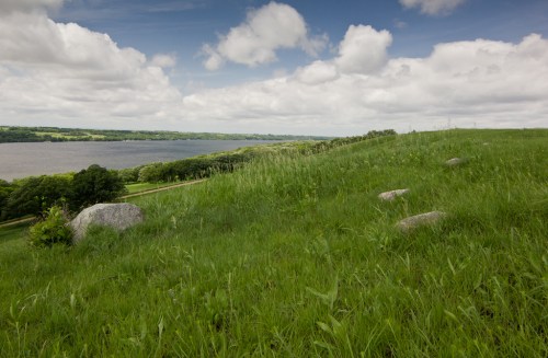 A grassy hillside with scattered rocks overlooks a calm river under a partly cloudy sky.
