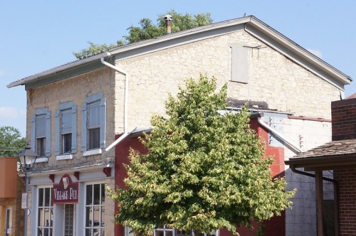 A two-story building with a stone exterior and blue shutters, next to a red building and a green tree in front.