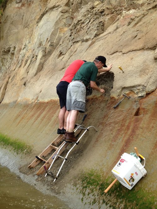 Two men on a ladder excavate a cliffside, using tools against a rocky surface near water. A bucket is nearby.