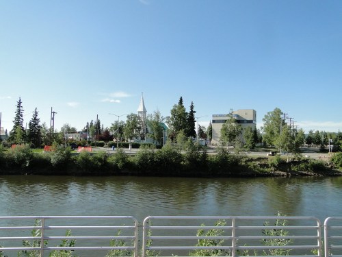 A calm river flows beside a tree-lined bank, with buildings and a church steeple visible in the background under a clear sky.