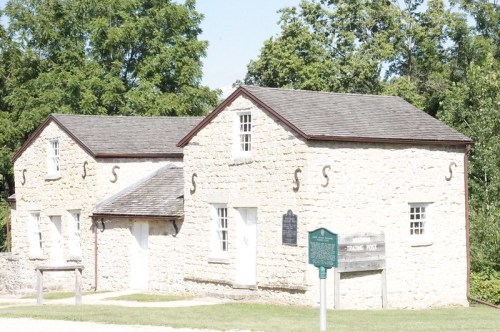 Historic stone building with a gabled roof, surrounded by trees and a grassy area, featuring a historical marker.