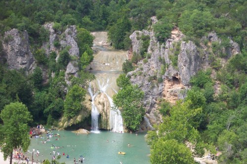 A scenic waterfall cascades into a turquoise pool, surrounded by lush greenery and rocky cliffs, with people enjoying the water.