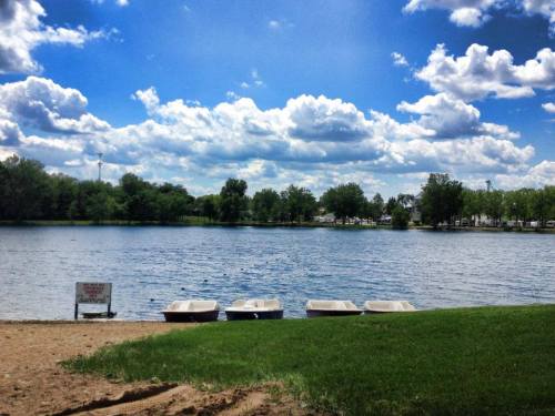 A serene lake scene with three boats on the shore, surrounded by green trees and a blue sky filled with fluffy clouds.