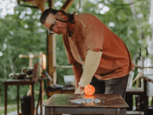 A glass artist shapes a glowing orange orb on a workbench in a workshop surrounded by greenery.